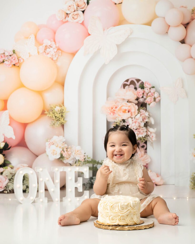 Dallas Butterfly Cake Smash Photography Featuring A Smiling Baby Girl Celebrating Her First Birthday With A Blush Pink Butterfly-Themed Cake Smash Setup And Floral Backdrop.