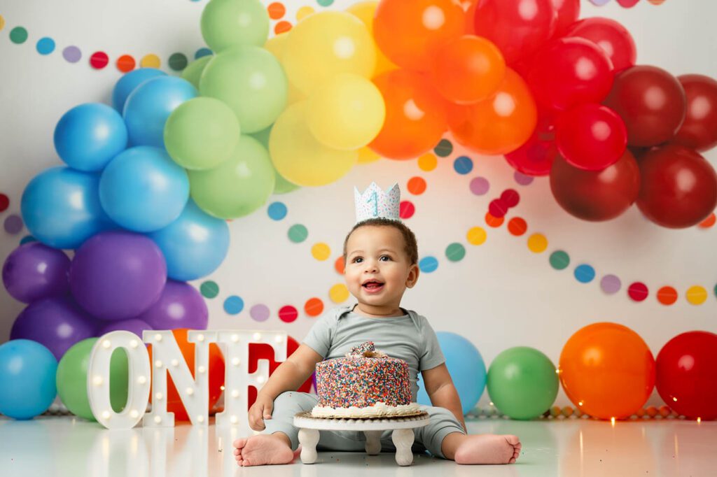 Dallas Rainbow First Birthday Cake Smash Photography Featuring A Baby Boy Wearing A Birthday Crown With A Sprinkle Cake, Rainbow Balloon Garland, And Colorful First Birthday Backdrop