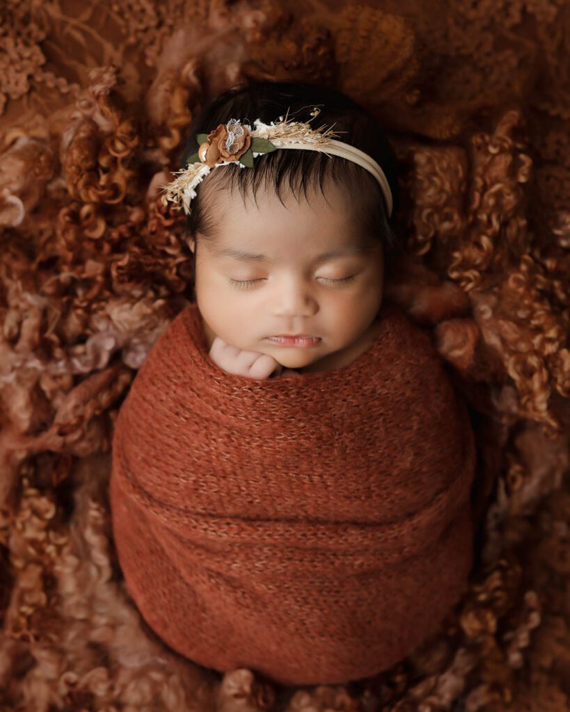 Sleeping Newborn Wrapped In A Rust-Colored Swaddle With Soft Professional Studio Lighting During A Dallas Newborn Photography Session.
