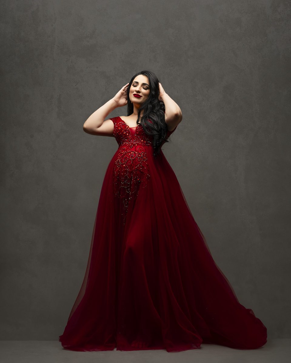 A woman in an elegant red gown posing with her hands in her hair against a gray backdrop.