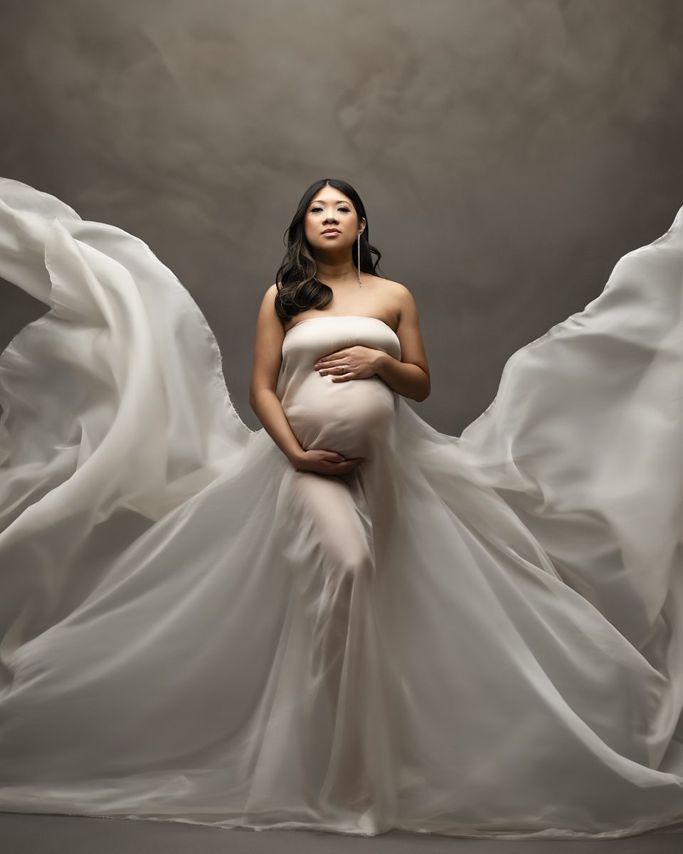 A pregnant woman in a white gown posing in front of a gray background.