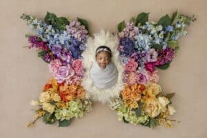 A Newborn Wrapped In A White Cloth Sleeping Amid A Colorful Floral Arrangement Shaped Like Butterfly Wings, Captured In Fine Art Newborn Photography