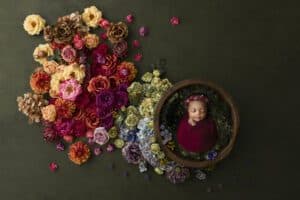 A Sleeping Baby Wrapped In A Red Blanket, Nestled In A Rustic Bowl Surrounded By A Colorful Array Of Flowers On A Dark Green Background, Captured In Fine Art Newborn Photography.