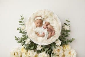 Newborn Baby Sleeping In A Round White Basket Surrounded By A Wreath Of White And Green Flowers During A Budget For A Newborn Photoshoot.