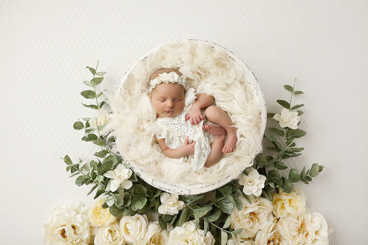 Newborn Baby Sleeping In A Round White Basket Surrounded By A Wreath Of White And Green Flowers During A Budget For A Newborn Photoshoot.
