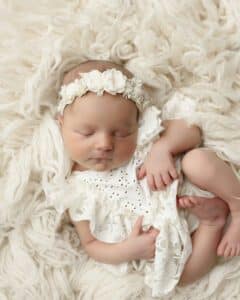 Newborn Baby Asleep On A Soft, Textured Fur During Their First Newborns First Photoshoot, Wearing A Delicate White Dress And A Floral Headband Newborn Photography Prop