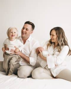 Father Holding Smiling Toddler While Mother Cradles A Newborn Baby, All Dressed In Light-Colored Clothing Beautifully Captured By The Role Of A Newborn Photographer