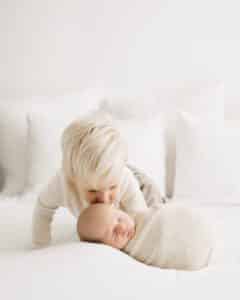 A Toddler Gently Kisses A Newborn Baby Wrapped In A Beige Blanket, Creating A Touching Moment Perfect For Capturing, Role Of A Newborn Photographer