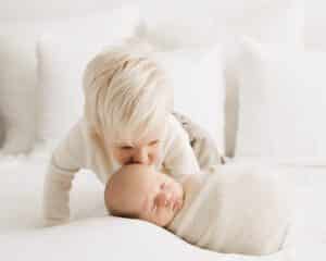 Older Brother Gently Kisses The Forehead Of A Swaddled Newborn Baby Lying On A Bed With White Pillows, Capturing Photography For Newborns