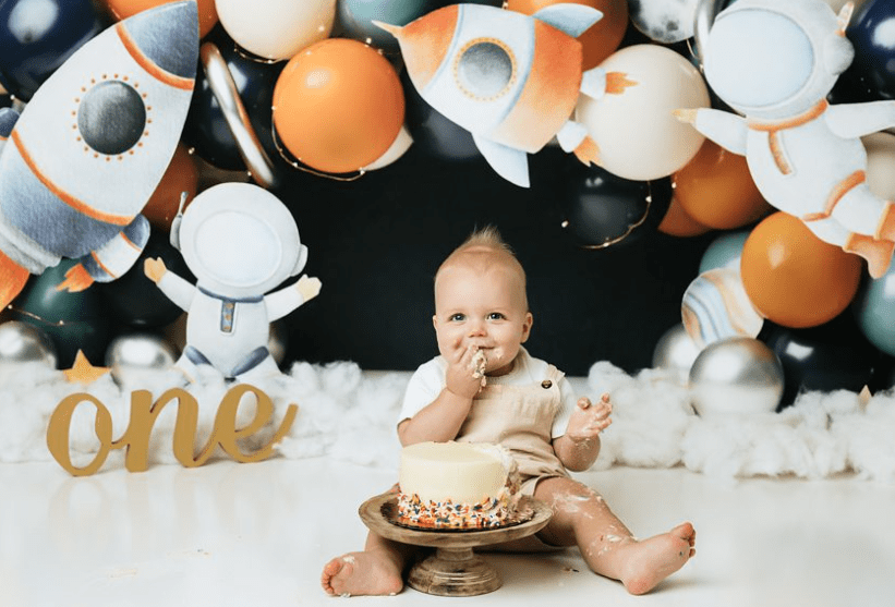 A Baby Sits On The Floor, Eating Cake With His Hands During A Delightful Cake Smash Session. The Background Is Decorated With Balloons And Space-Themed Cutouts. The Word &Quot;One&Quot; Is Displayed, Indicating A First Birthday Celebration Captured In Charming Cake Smash Photography.