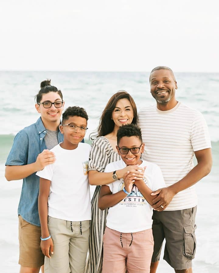 A family of five poses together on a beach. The group consists of two adults and three children. The ocean is visible in the background.