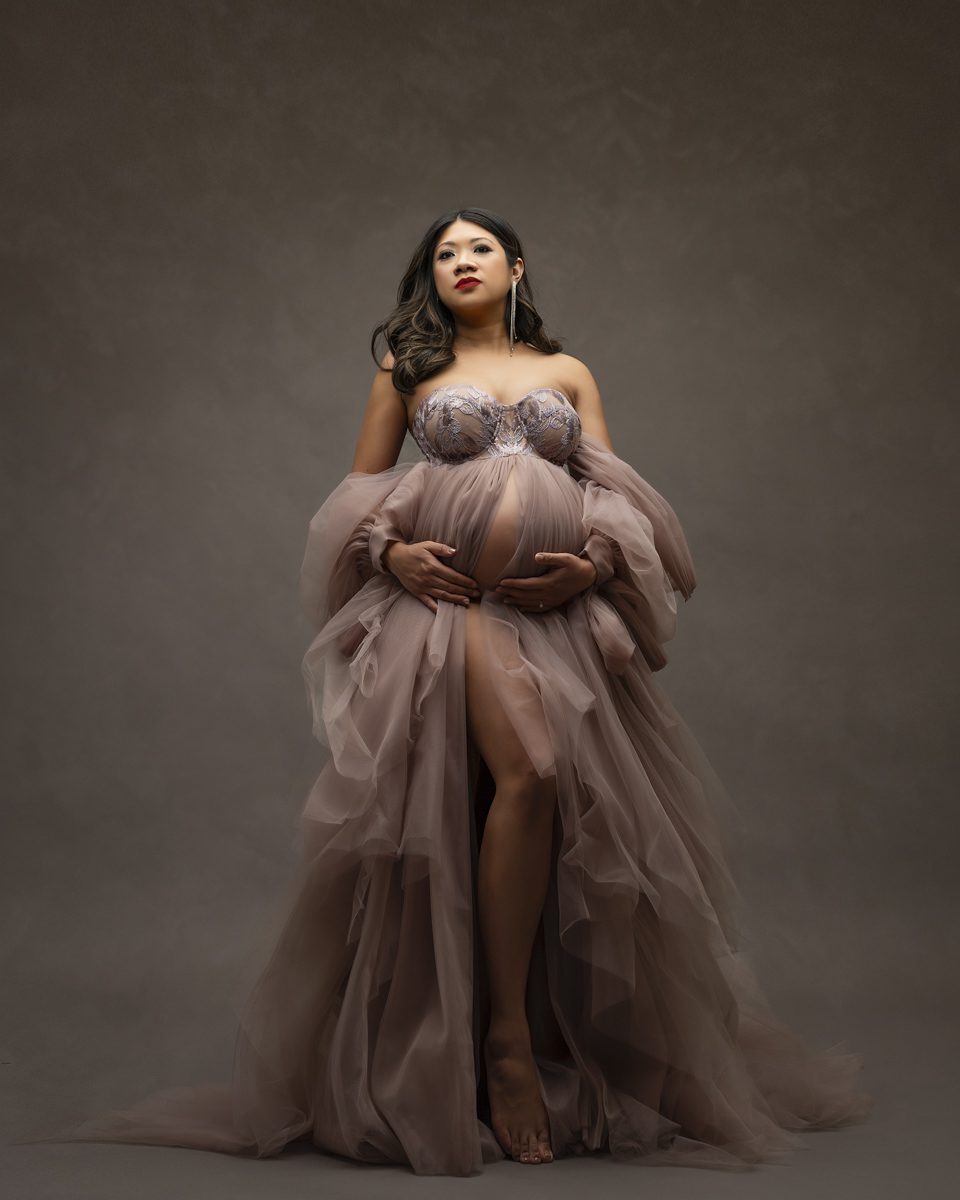 Pregnant woman in an elegant mauve maternity photography gown holding her bump, standing against a grey backdrop.