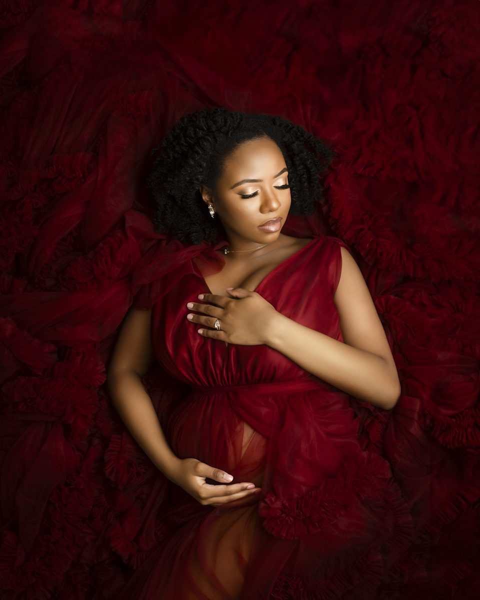 A pregnant woman with dark hair, wearing a flowing red dress, gently holds her belly while surrounded by rich red fabric for her maternity photographer