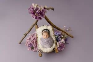 A Sleeping Baby, Wrapped In A Purple Blanket And Headband, Lies On A Small Wooden Bed With A White Fur Cover. Surrounding The Bed Are Flowers And Branches, All Placed On A Light Purple Background. This Enchanting Scene Showcases The Artistry Of Dallas Newborn Photography.