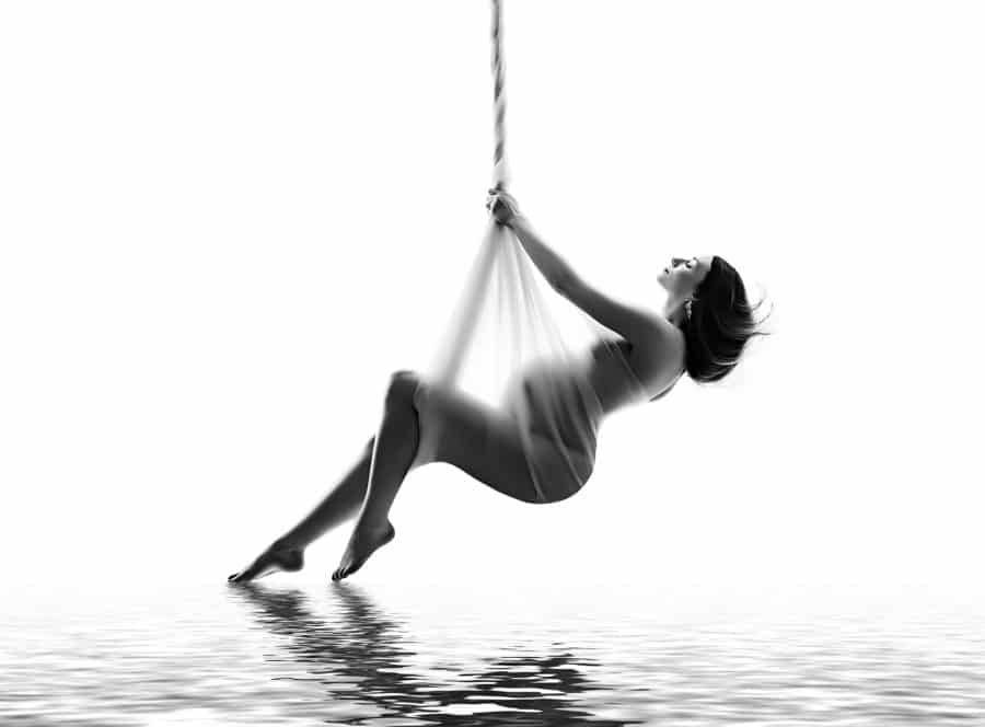 A woman swings on a rope above water in a black-and-white maternity photoshoot in dallas