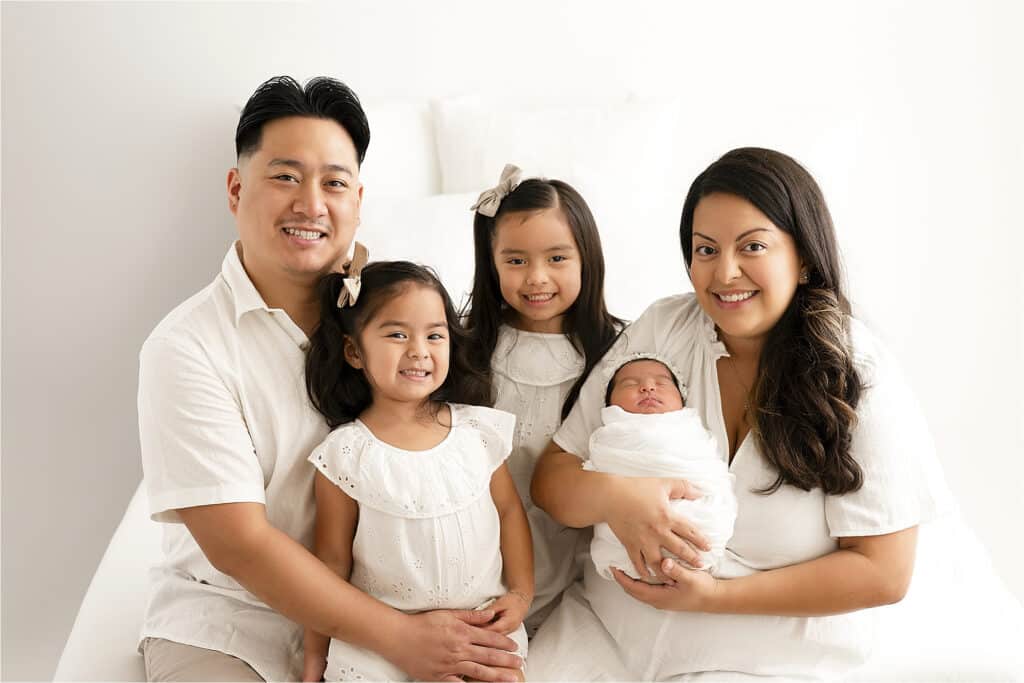 A Family Of Five, Including A Man, Two Young Girls, And A Woman Holding A Swaddled Baby, All Dressed In White, Posed Together In A Well-Lit Setting For Their Lifestyle Newborn Pictures In Dallas, Tx.