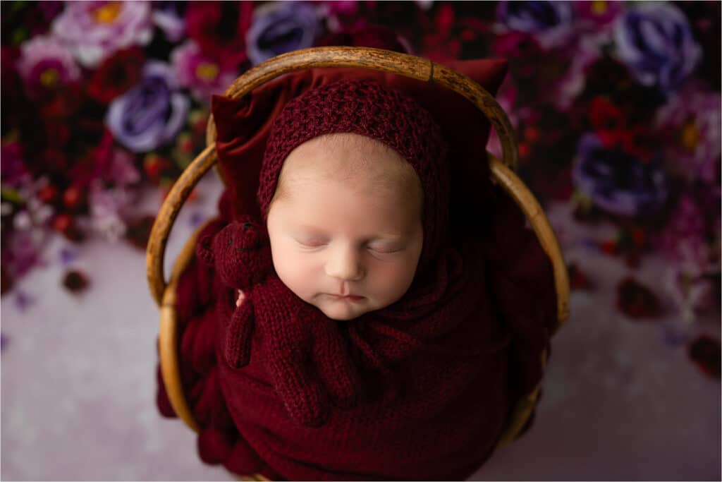 A Newborn Baby Is Swaddled In A Deep Red Knit Blanket And Hat, Sleeping Peacefully In A Wicker Basket Against A Backdrop Of Dark Red And Purple Flowers. Capturing Moments Like These Requires The Best Lens For Newborn Photography To Ensure Every Detail Is Perfect.