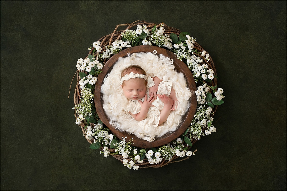 A Newborn Baby Lies In A Brown, Bowl-Shaped Basket, Surrounded By White Flowers And Green Leaves On A Dark Background. The Baby Is Wrapped In A Soft White Blanket And Wears A Matching Headband. Capturing This Precious Moment With The Best Lens Is Essential For Stunning Newborn Photography In Dallas.