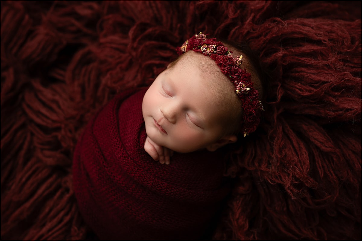 A Newborn Baby Wrapped In A Burgundy Cloth Sleeps On A Textured Burgundy Blanket, Wearing A Matching Floral Headband—A Perfect Capture Of Dallas Newborn Photography Through The Lens.