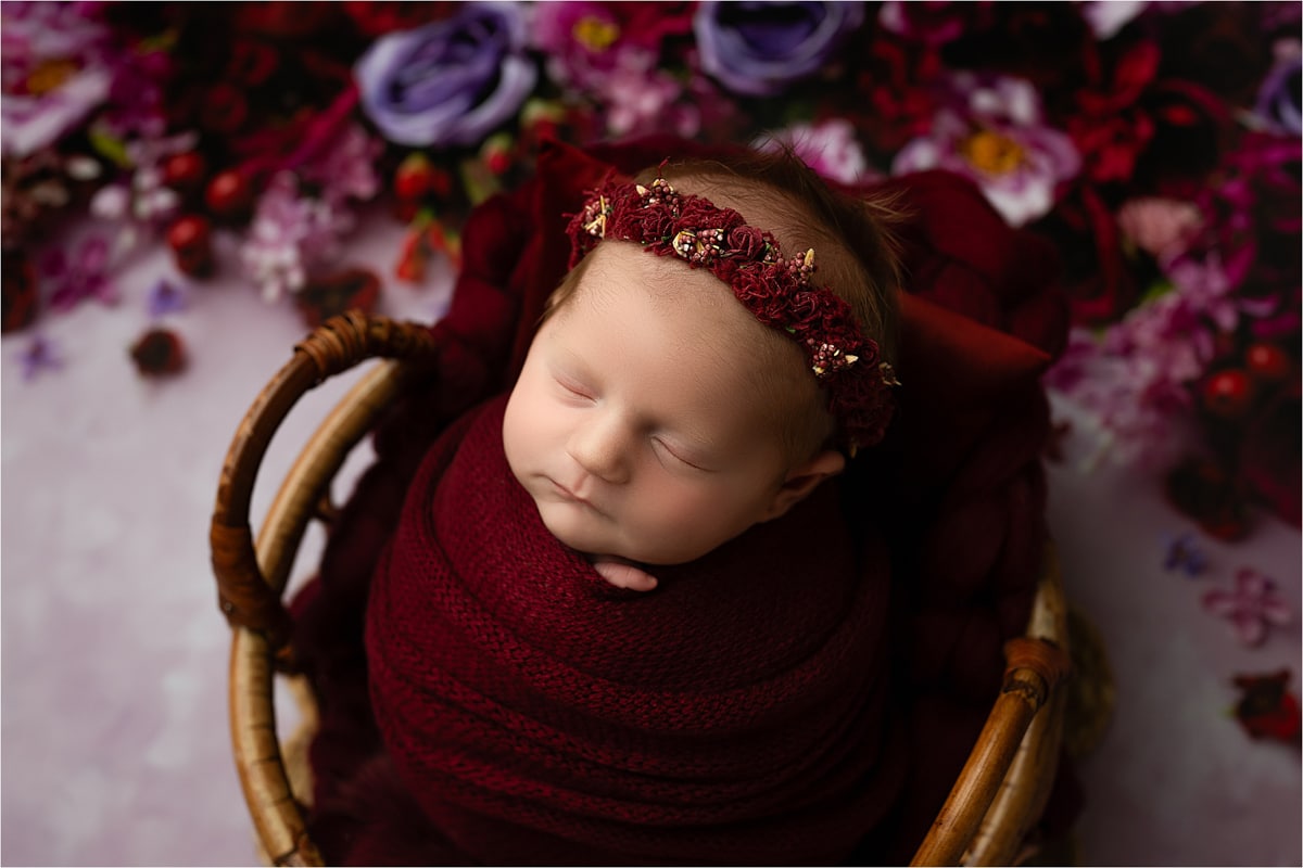 In The Heart Of Dallas, A Newborn Baby, Wrapped In Burgundy Fabric With A Matching Headband, Sleeps Peacefully In A Basket Surrounded By A Floral Background—An Exquisite Moment Of Newborn Photography.