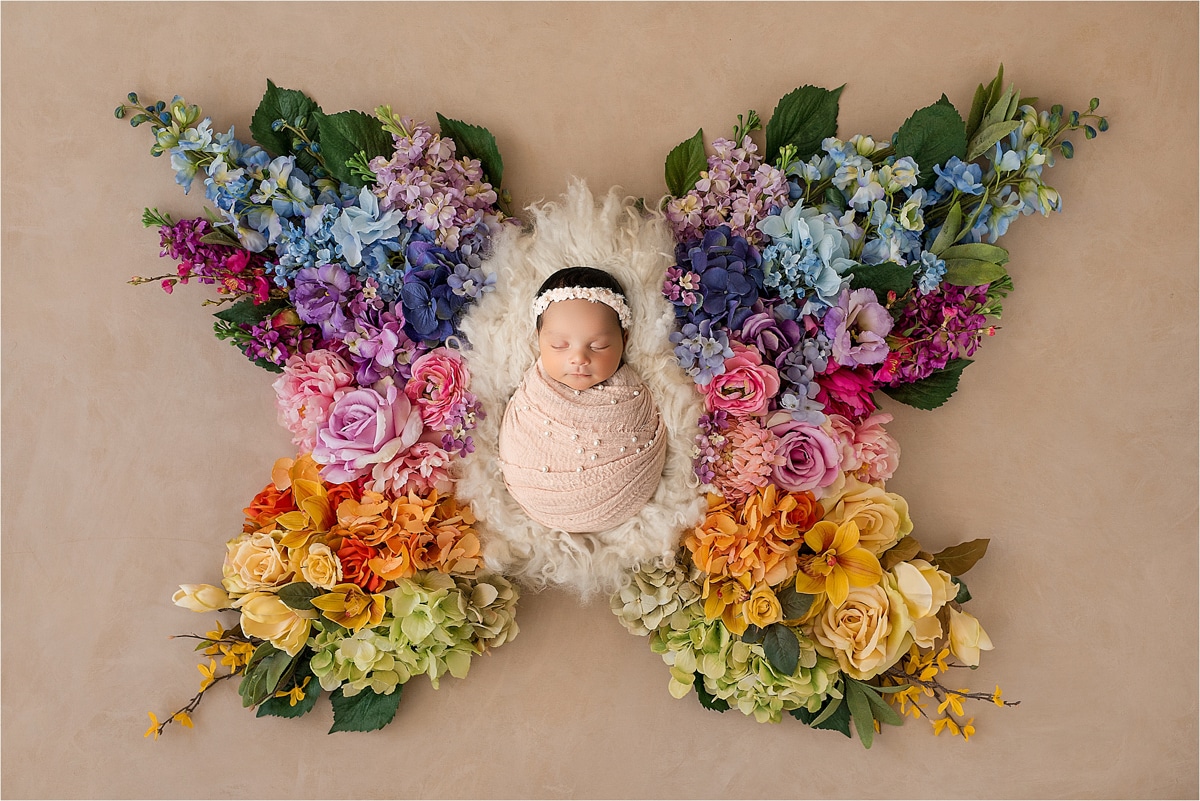 A Baby Wrapped In A Pink Blanket Sleeps On A Furry White Blanket, Surrounded By Colorful Flowers Arranged In The Shape Of Butterfly Wings—A Precious Moment Captured Perfectly By Dallas Tx Newborn Photography.