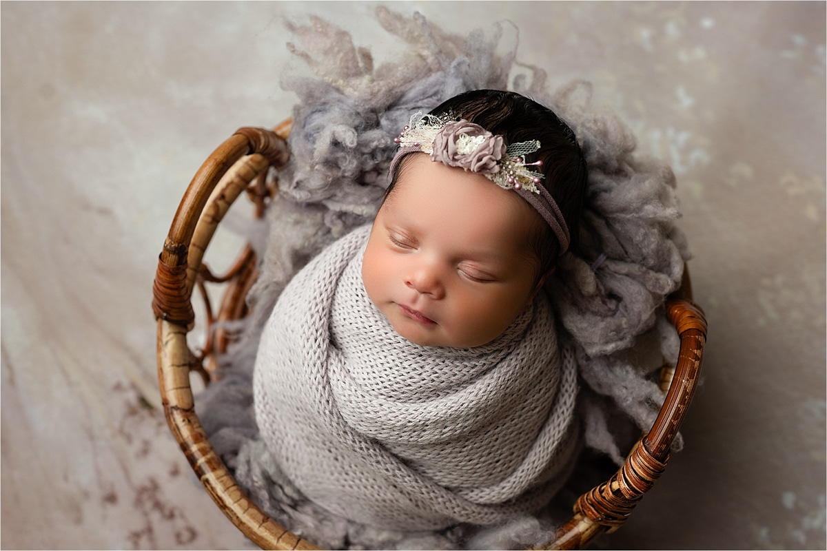 A Newborn Baby Is Swaddled In A Knitted Wrap And Lying In A Wicker Basket, Wearing A Floral Headband. The Soft Background Complements The Cozy Setting, Capturing The Essence Of Newborn Photography. For Those In Dallas Tx, Prices And Packages Are Available Upon Inquiry.