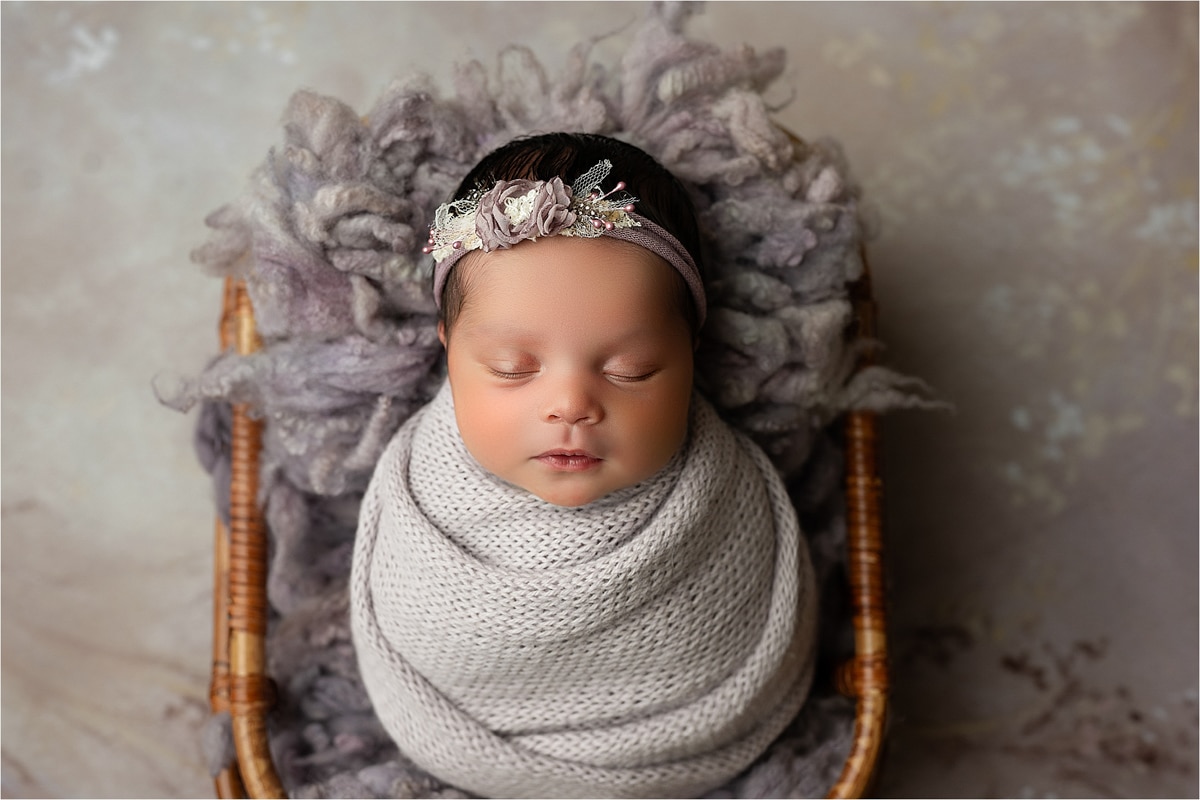 A Newborn Baby Is Swaddled In A Gray Knit Blanket, Resting In A Small Wooden Basket, Wearing A Decorative Headband. For Those In Dallas Tx Seeking Professional Newborn Photography, Inquire About Our Affordable Prices For Capturing These Precious Moments.