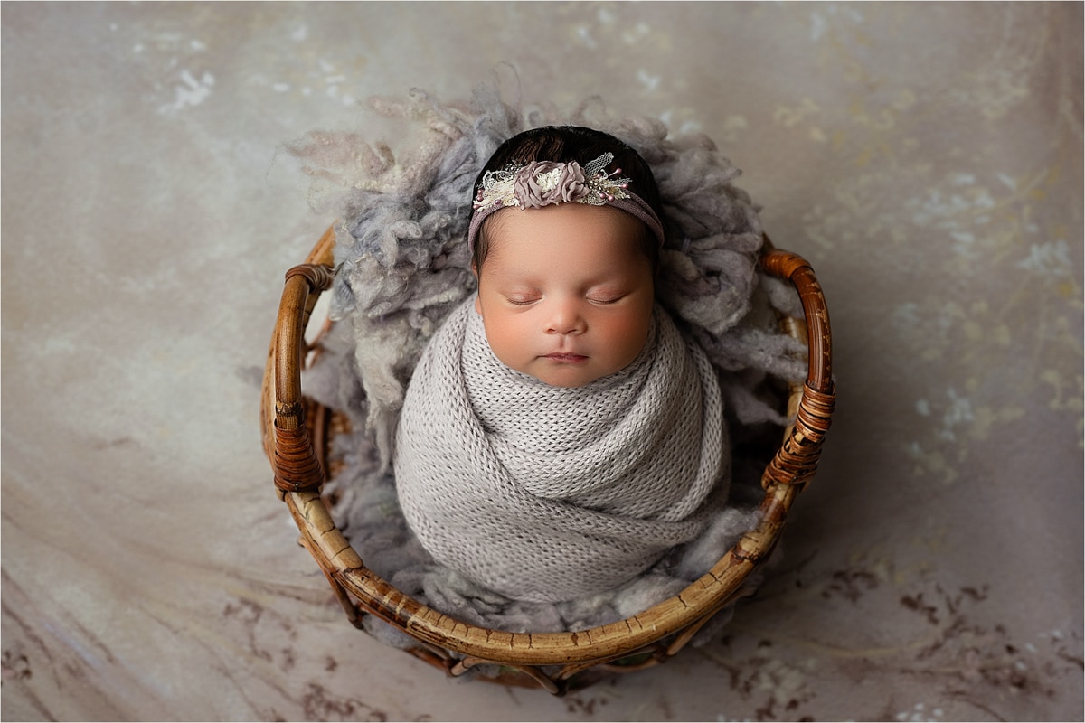 A Newborn Baby Is Swaddled In A Knit Blanket, Lying In A Wicker Basket With Soft, Fluffy Padding, And Wearing A Floral Headband. Capture This Precious Moment With Our Affordable Newborn Photography Prices In Dallas Tx.