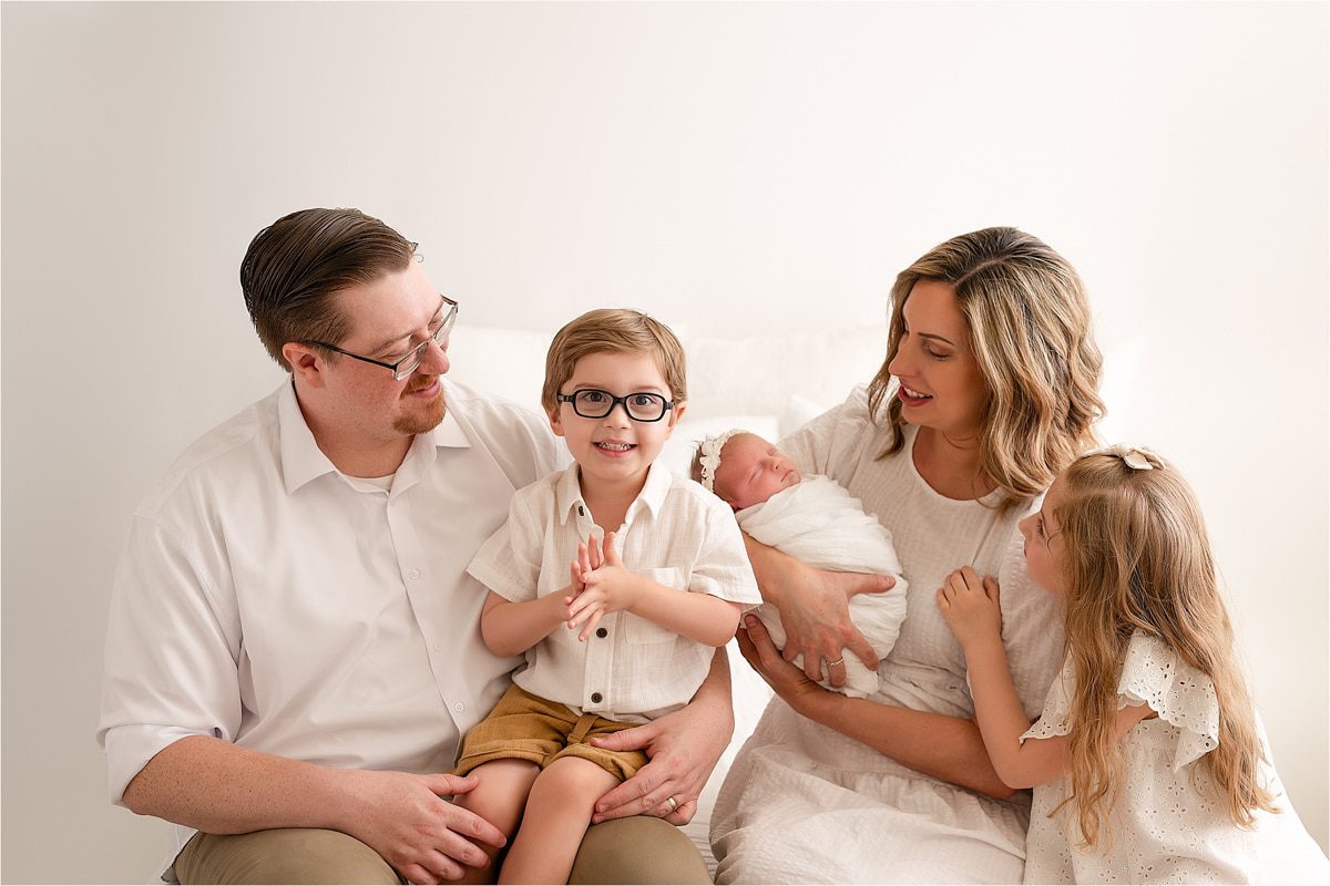 A Family Of Five Poses For A Photo Taken By Their Newborn Photographer. The Parents Sit At Either Side, With The Father Holding The Young Boy And The Mother Holding An Infant Wrapped In A White Blanket. The Daughter Looks At The Baby.