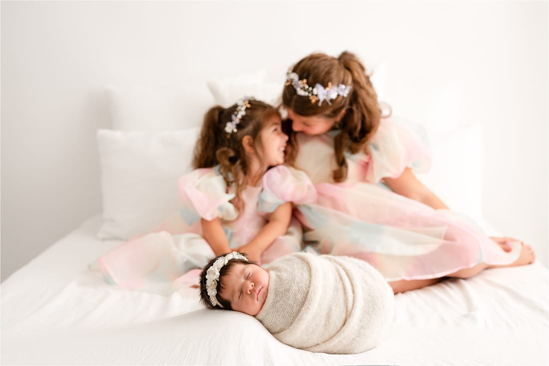 Three Young Children: An Infant Swaddled In A Blanket In The Foreground, And Two Young Girls Wearing Pastel-Colored Dresses And Floral Headbands Sitting Together In The Background On A White Bed, Capture A Perfect Moment That Showcases The Artistry Of Newborn Photographers.