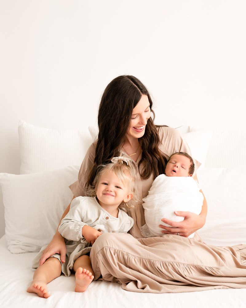 Mom And Daughter Sitting On A White Bed With Newborn During A Newborn Photoshoot 