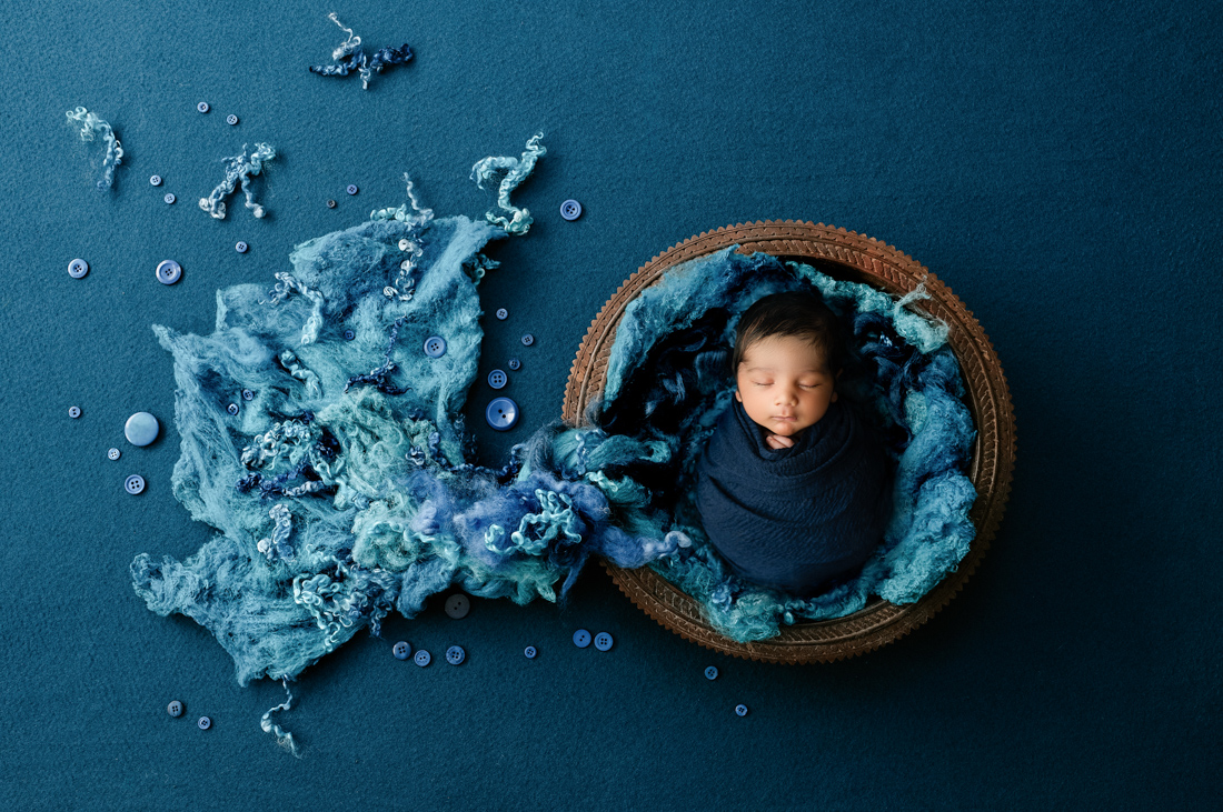 sleeping newborn posed in a bowl surrounded by blue fabric for newborn photographer dallas