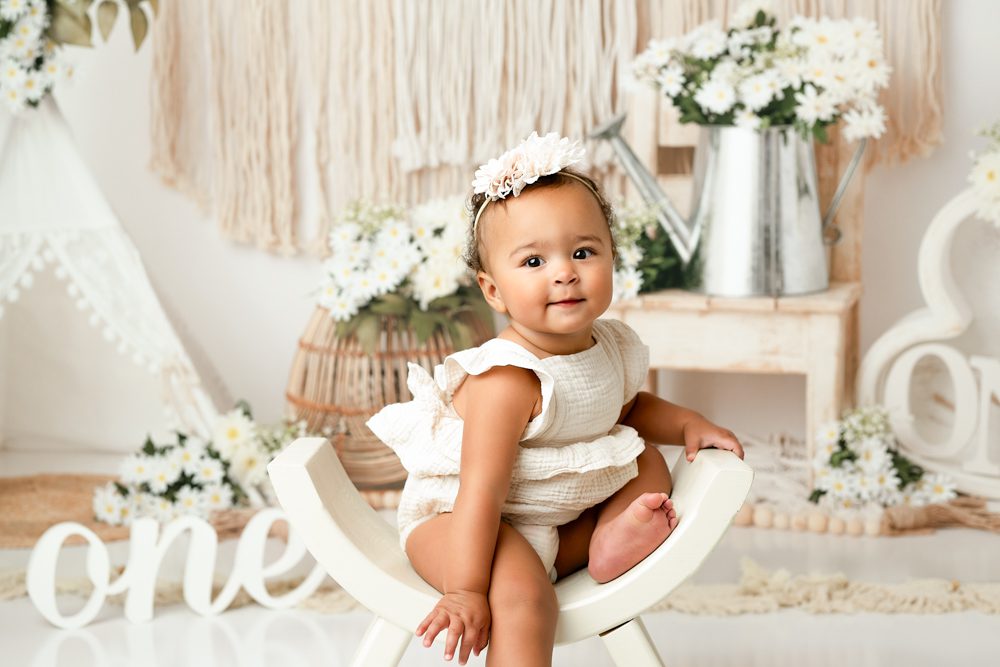 A Baby Wearing A Flower Headband Sits On A White Curved Chair, Surrounded By White Daisies And Rustic Decor.