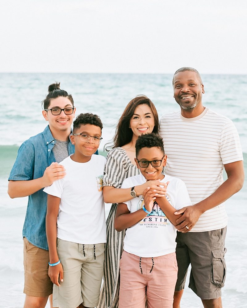 A family of five poses on a beach. The background shows the ocean and a clear sky.