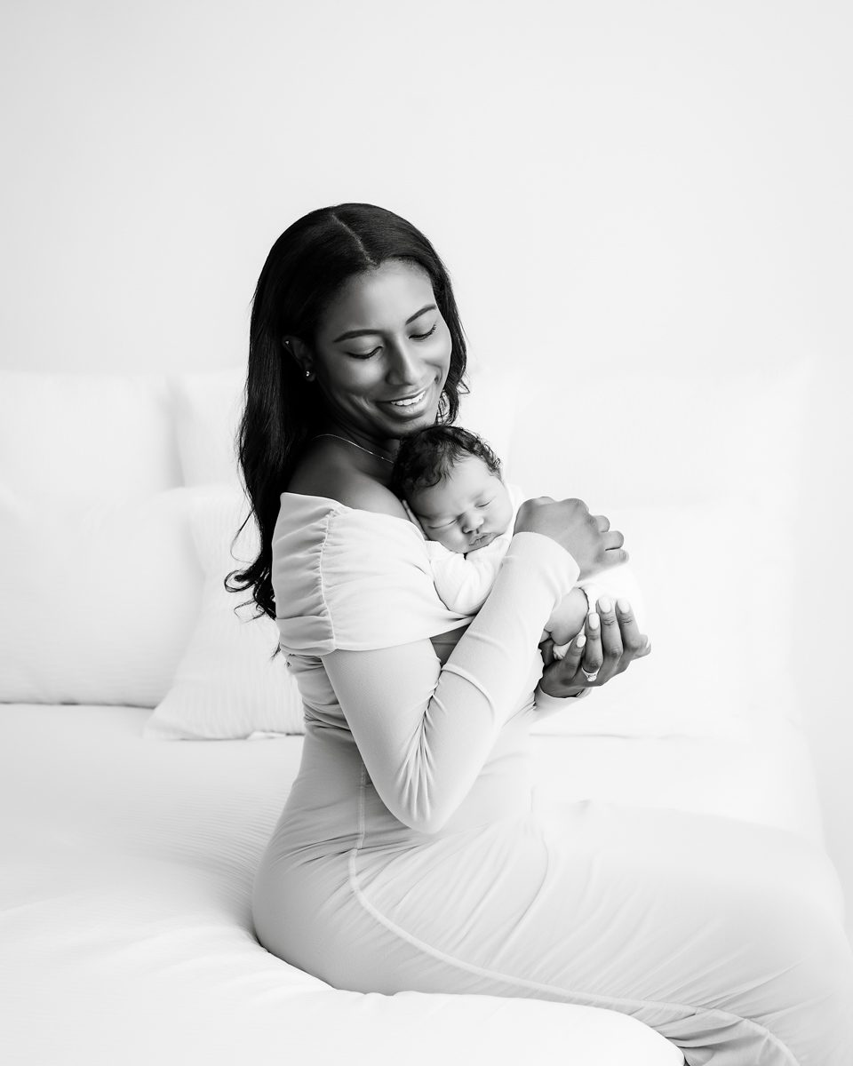 Mother Sitting Smiling On Bed Cradling Her Sleeping Newborn For Dallas Fine Art Newborn Photographer
