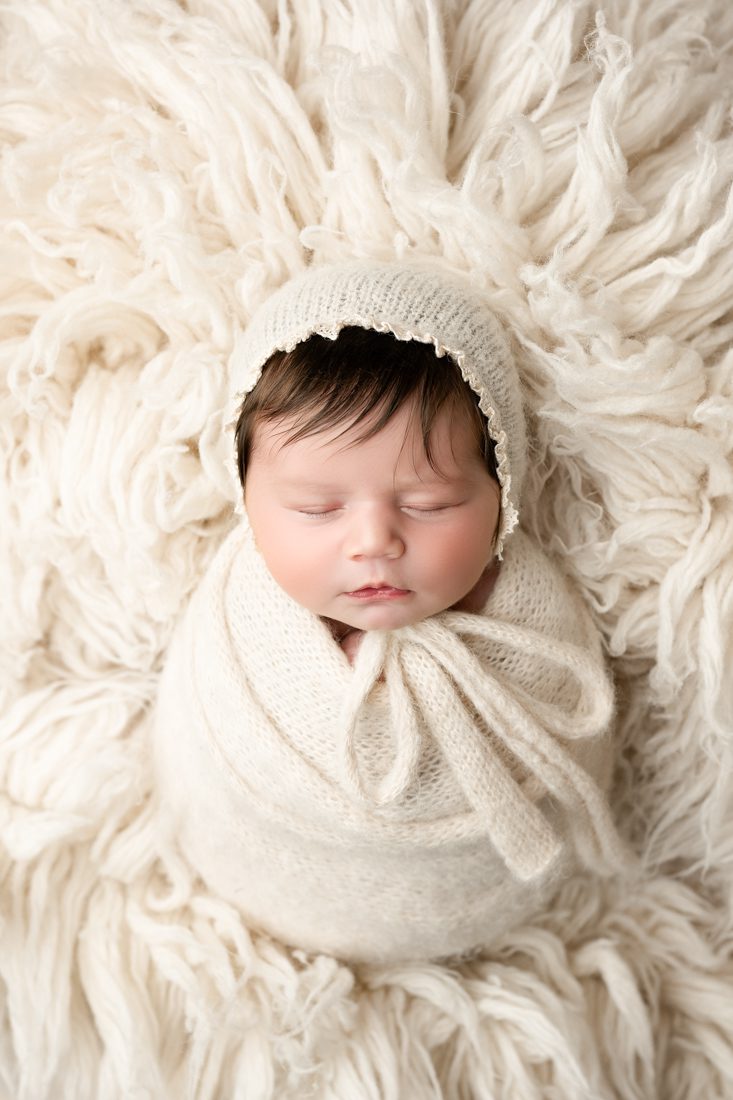 A Newborn Photographer In Dallas Captures A Baby With Dark Hair Sleeping Peacefully, Wrapped In A Cream-Colored Knit Blanket And Bonnet, Lying On A Soft, Textured Cream Background.