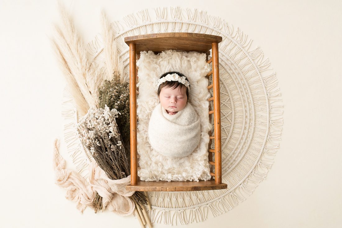 A Newborn Baby Wrapped In A Cream Blanket Lies In A Small Wooden Bed, Surrounded By Dried Flowers And Pampas Grass—A Perfect Scene For Those Seeking Newborn Photographer Tips Or Inspiration For Stunning Newborn Photography.