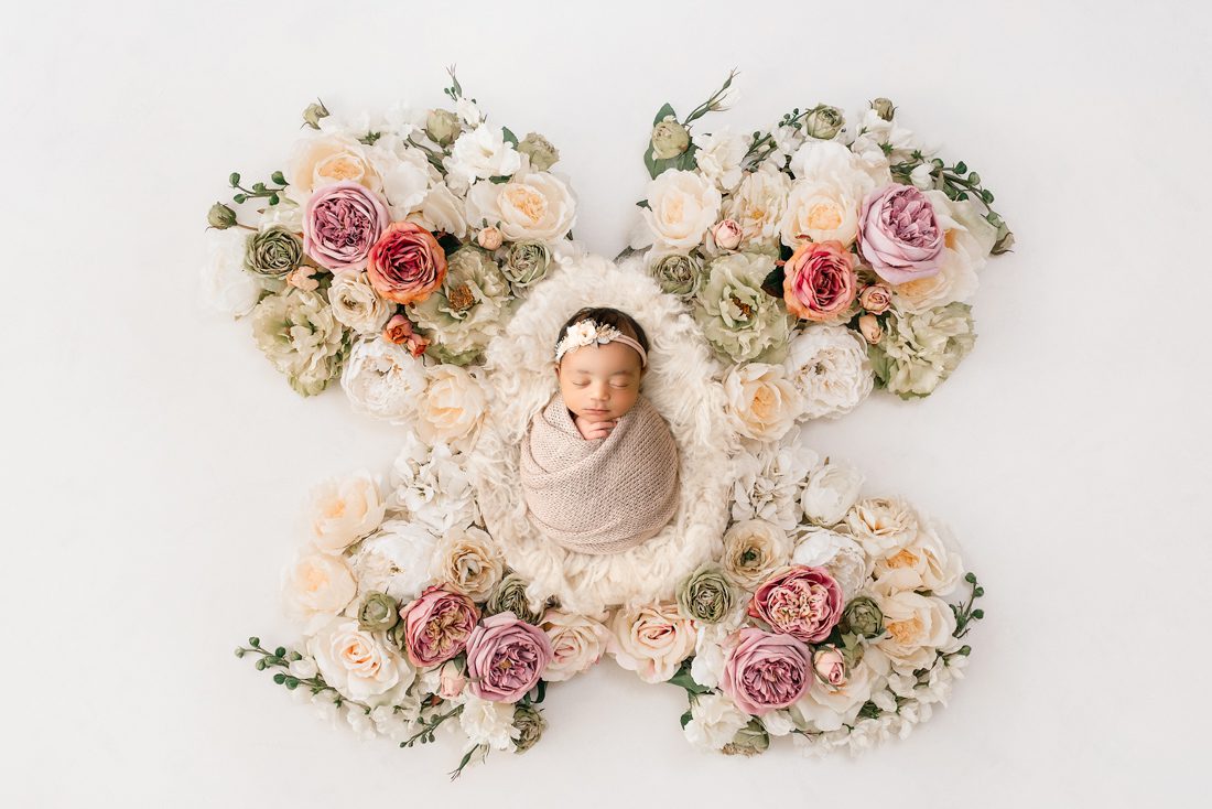 Newborn Baby Wrapped In A Beige Blanket Lies On A White Surface, Surrounded By Flowers Arranged In The Shape Of Butterfly Wings—A Dreamy Scene Perfect For Newborn Photography.