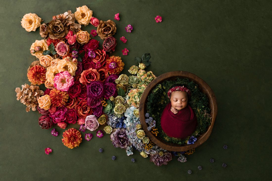 A Newborn Baby Wrapped In A Burgundy Blanket Sleeps In A Wooden Bowl, Surrounded By Colorful Artificial Flowers On A Green Background—Perfect For Showcasing The Beauty Of Newborn Photography And Dispelling Common Newborn Photography Myths.