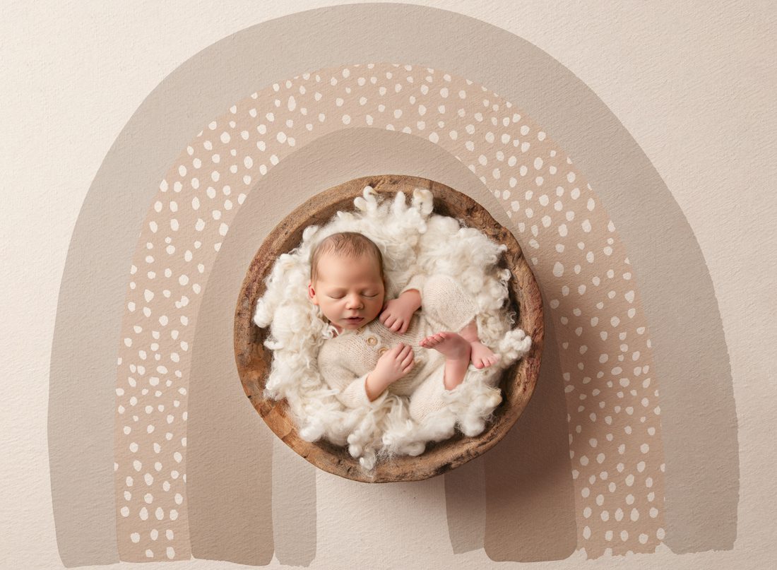 Newborn Photography: A Newborn Baby Sleeps In A Wooden Bowl Filled With Soft White Wool, Set Against A Beige Background With A Stylized Arch And Dotted Pattern.