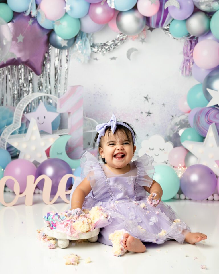 baby in a lavender dress with a matching bow sits on the floor smiling for cake smash photographer dallas