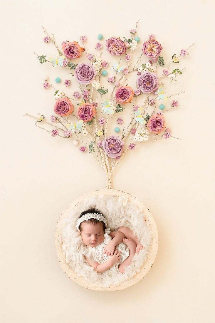 A Newborn Baby Sleeps In A Fluffy White Basket, Beneath Pink And Purple Artificial Flowers Arranged Like A Tree—A Dreamy Setup Perfect For A Newborn Photography Mini Session.