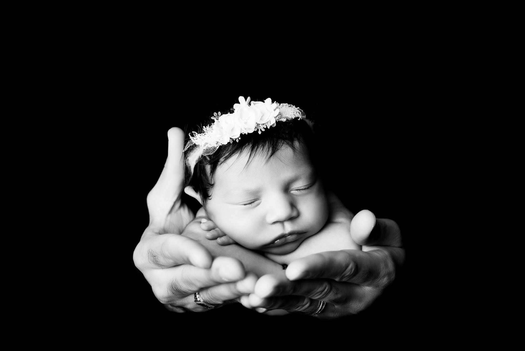 Black And White Newborn Portrait Of Baby Girl Wearing Floral Headband, Gently Cradled In Parents’ Hands – Dallas Newborn Photoshoot