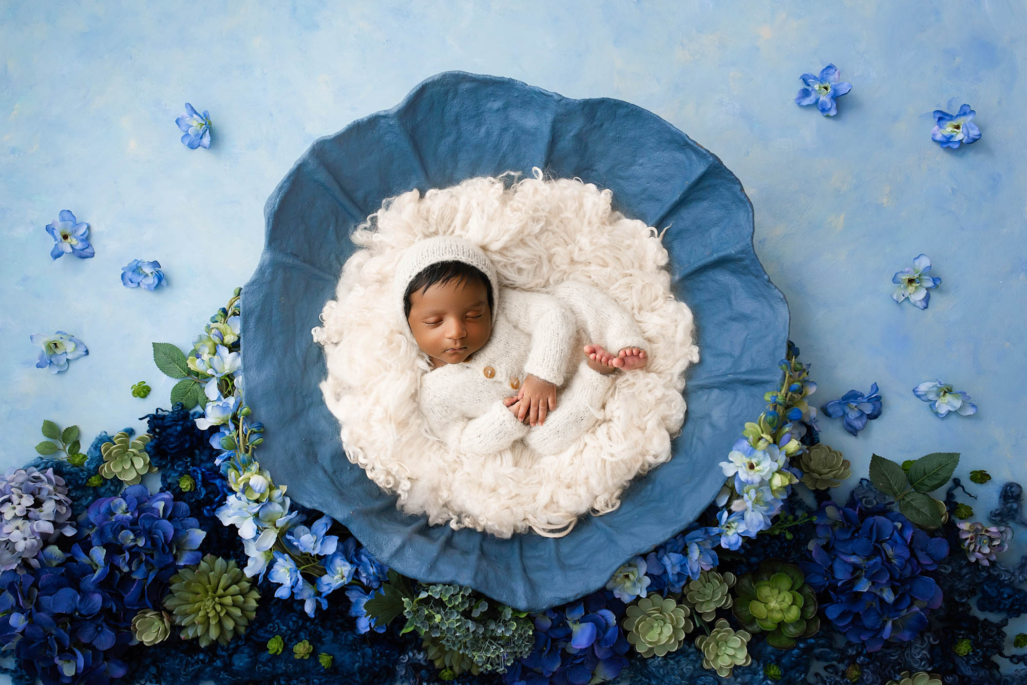 Newborn baby boy dressed in white knit outfit and bonnet, peacefully sleeping in a fluffy nest inside a blue flower surrounded by hydrangeas – Dallas newborn photographer