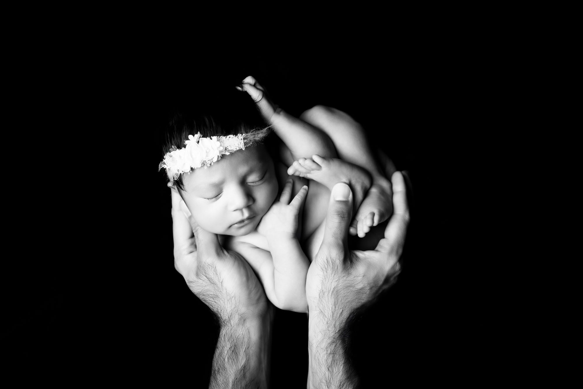 Fine Art Newborn Photography In Dallas – Baby Girl In Floral Headband Curled In Parents’ Hands, Black And White Portrait