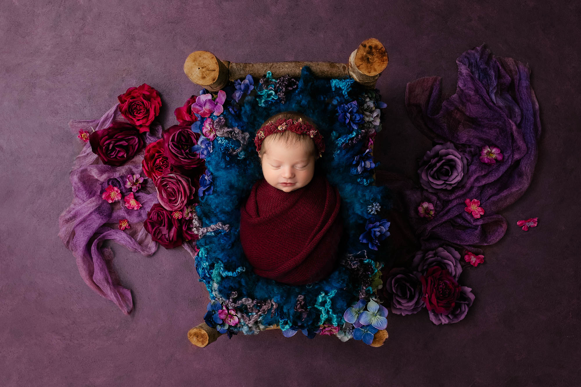 Newborn baby wrapped in burgundy swaddle on a rustic mini bed with purple and blue floral setup in Dallas photography studio for newborns first photo shoot