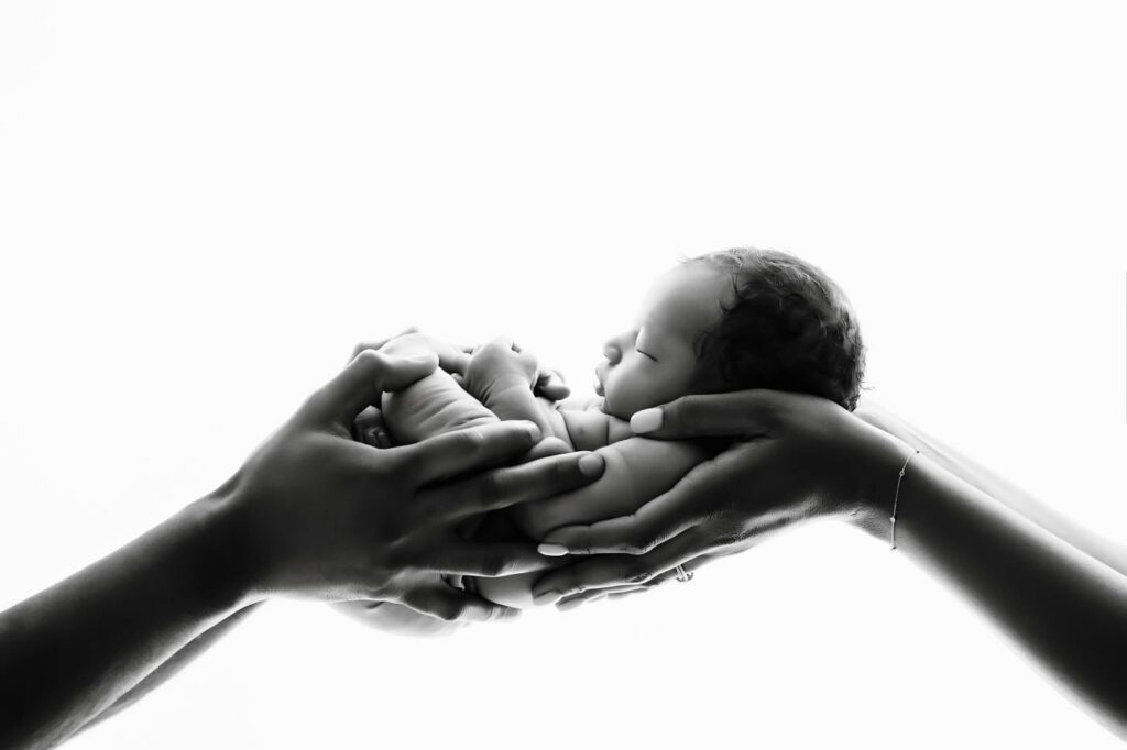 Black And White Newborn Portrait Featuring A Sleeping Baby Cradled In The Hands Of Loving Parents During A Dallas Newborn Photography Session