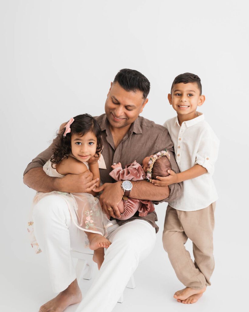 Father Holding His Newborn Daughter While Her Older Brother And Sister Gather Close During A Dallas Newborn Photography Session