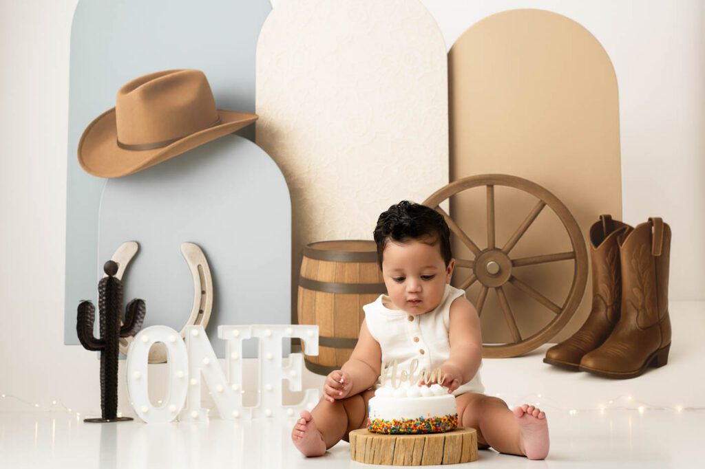What If Your Baby Won’t Smash The Cake During A Cake Smash Session? 15 Western Cake Smash Theme Created By A Dallas Cake Smash Photographer Featuring A Cowboy-Inspired First Birthday Setup With Rustic Props, A Birthday Cake, And A Little Cowboy Exploring The Frosting.