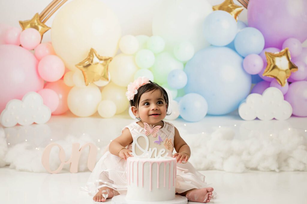 One-Year-Old Baby Girl Celebrating Her First Birthday With A Custom Cake Smash Session In A Dallas Photography Studio Featuring A Pastel Rainbow Balloon Backdrop.